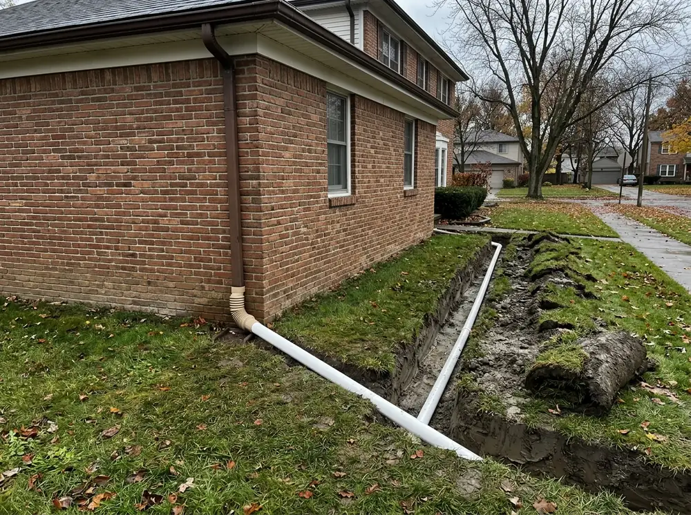 Trench across a lawn beside a brick house with a white rigid PVC pipe connected to the base of a downspout through an adapter, sloping away from the foundation